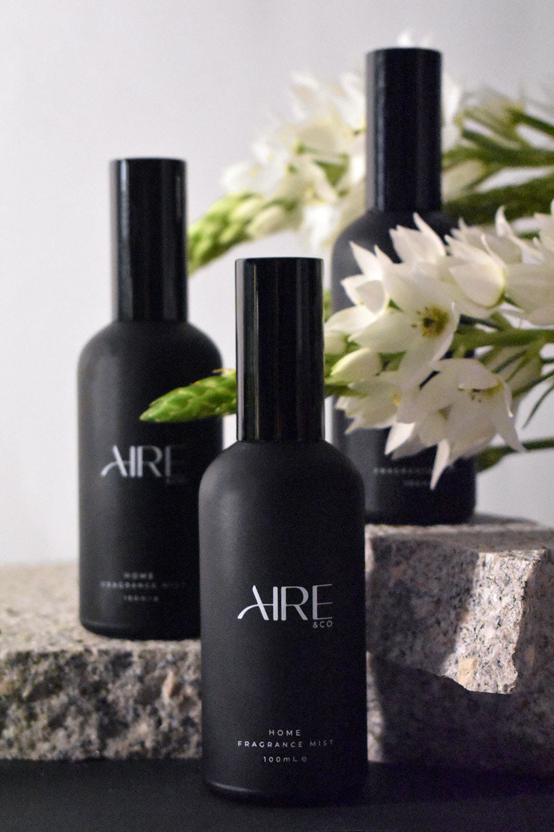 Three Bloom Aire & Co home fragrance mist bottles sitting grey rocks and with white flowers in the background