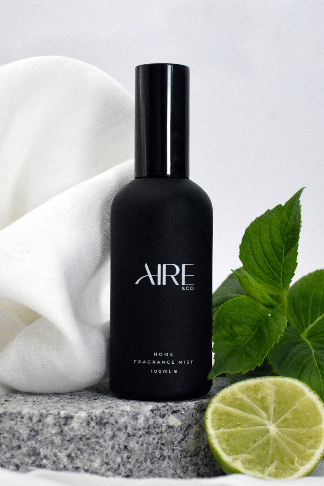 Linen Aire & Co home fragrance mist bottle with green leaves, white fabric and a lime on a gray stone.